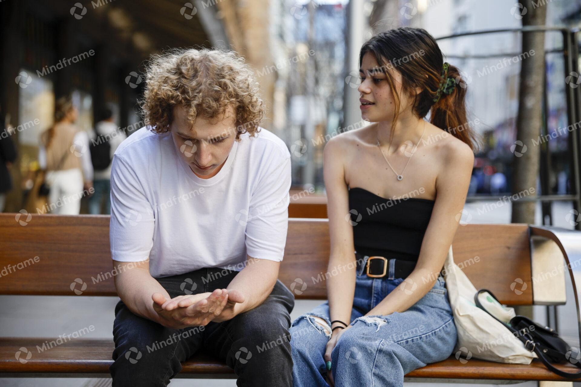 Young man with head bowed talking to young woman on city bench