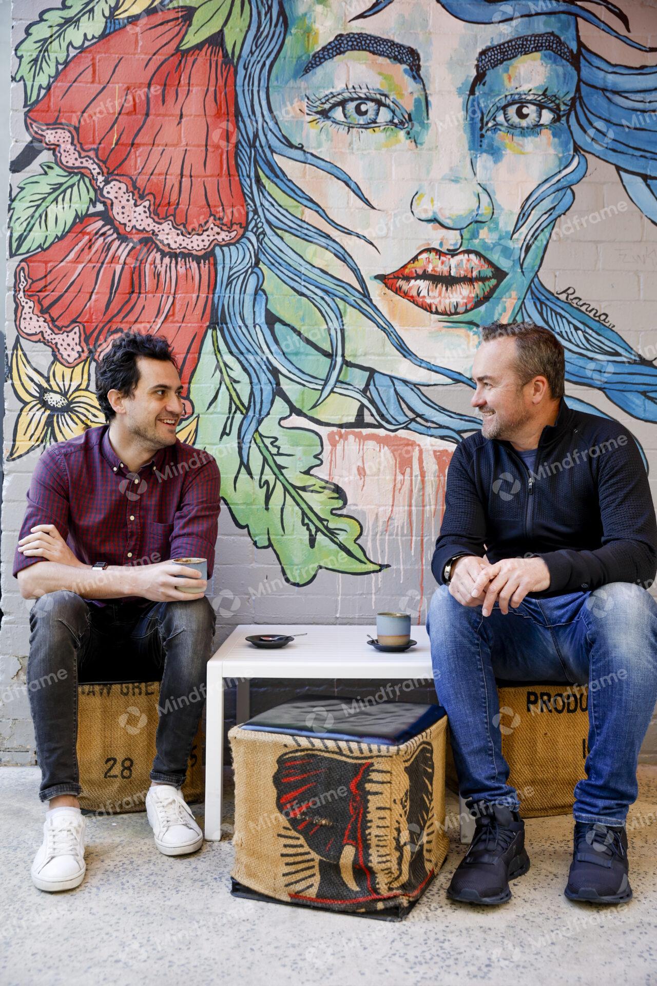 Two people sitting next to a mural talking with coffee
